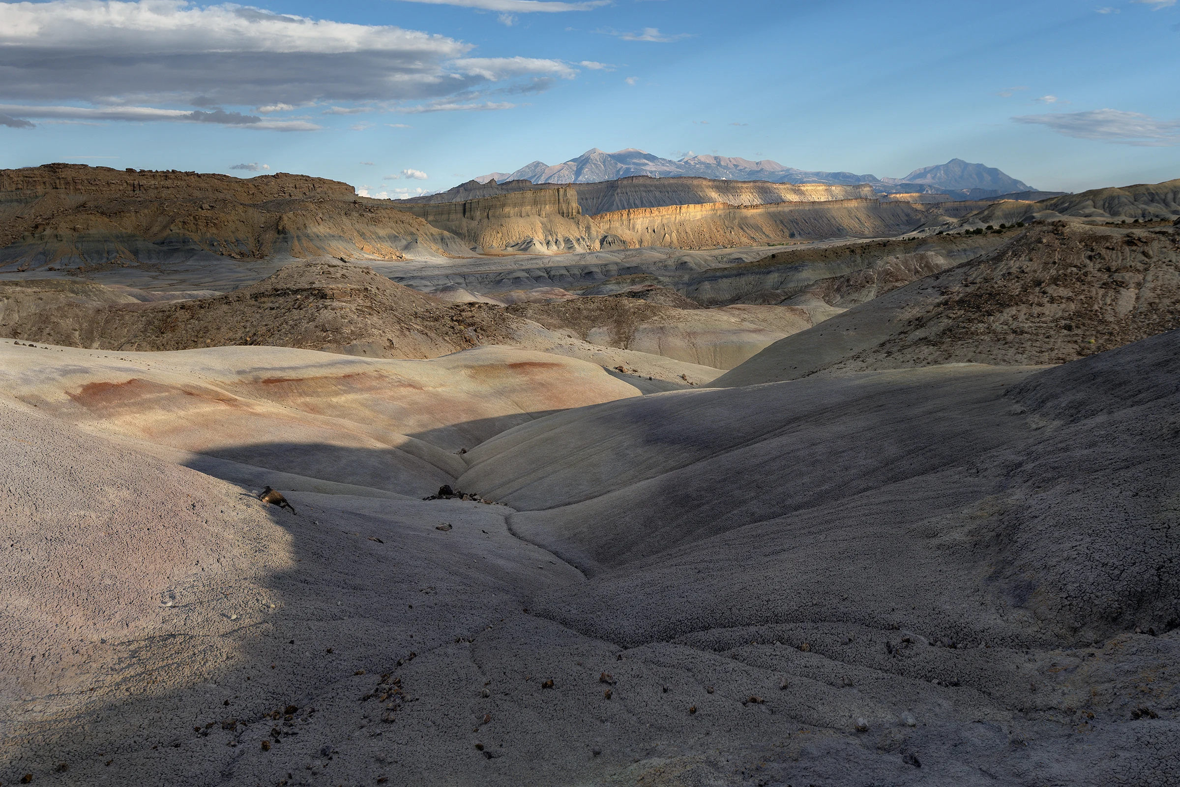 Rounded gray badland hills and eroded gullies lead toward layered cliffs and distant mountains, with patches of golden sunlight and rust-colored bands beneath a partly cloudy blue sky.