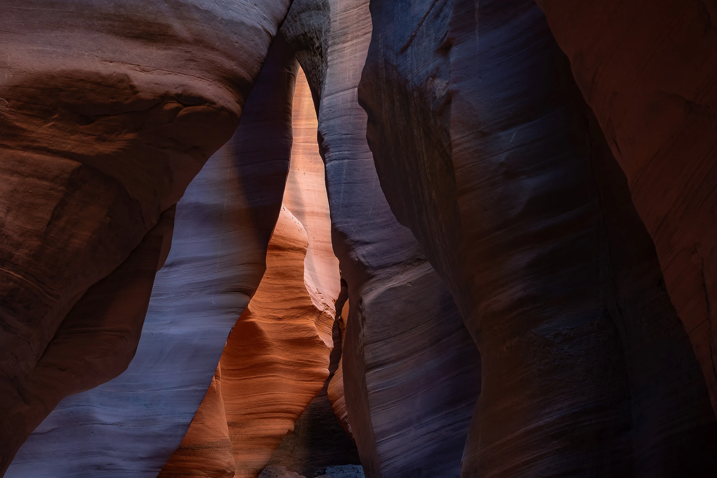 Curving sandstone walls enclose a narrow slot canyon, with warm orange light illuminating the central passage while reddish brown and purple shadows accentuate the surrounding rock’s flowing contours and fine horizontal layers.