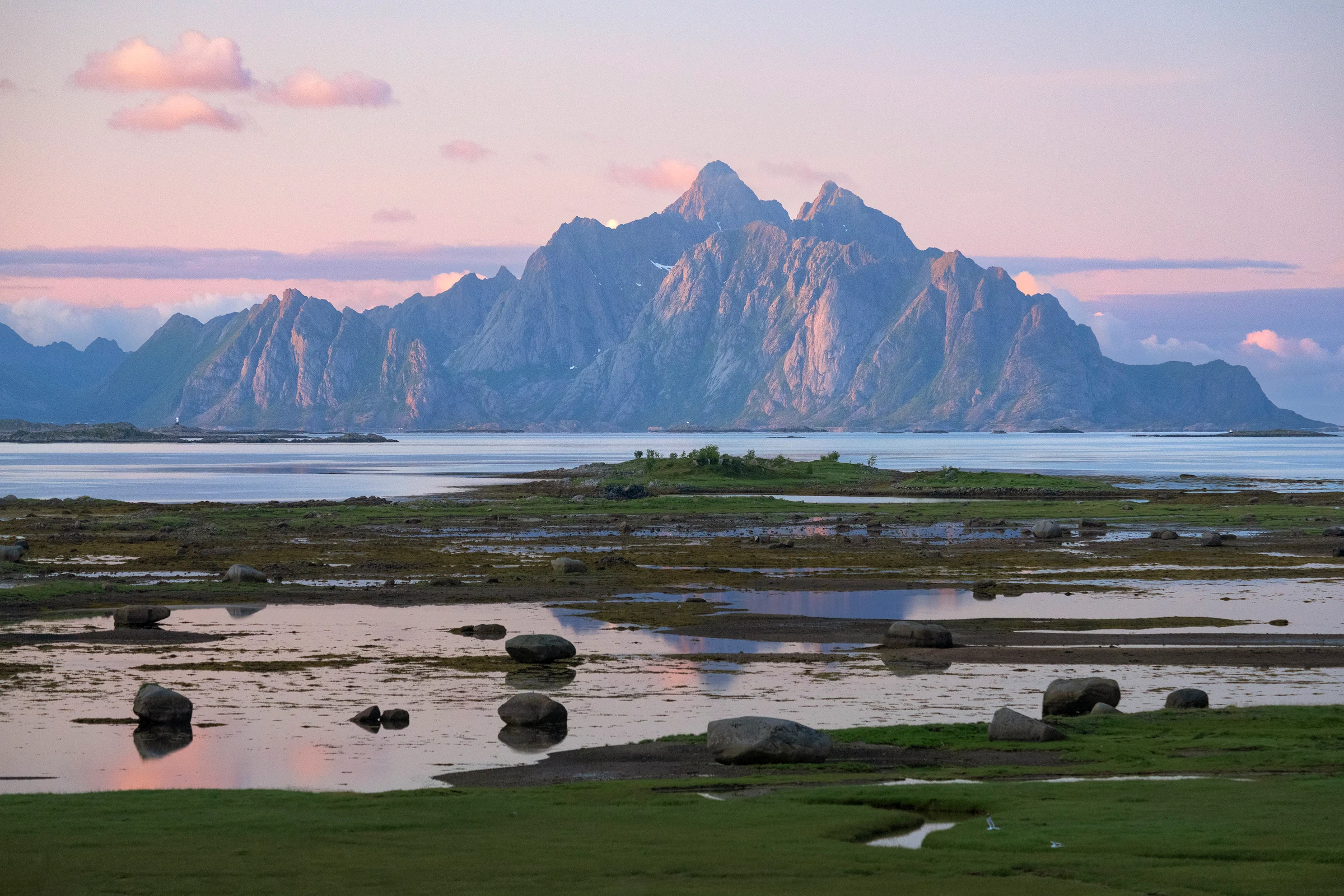 A distant towering mountain with many peaks rises out of the ocean during a sunset of purple and pink colors
