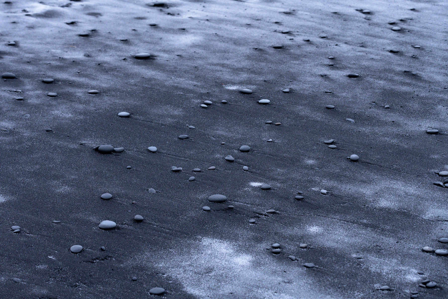 Smooth gray pebbles lie scattered across dark sand, with pale streaks and shallow diagonal grooves forming subtle patterns in a close, downward view tinted blue.
