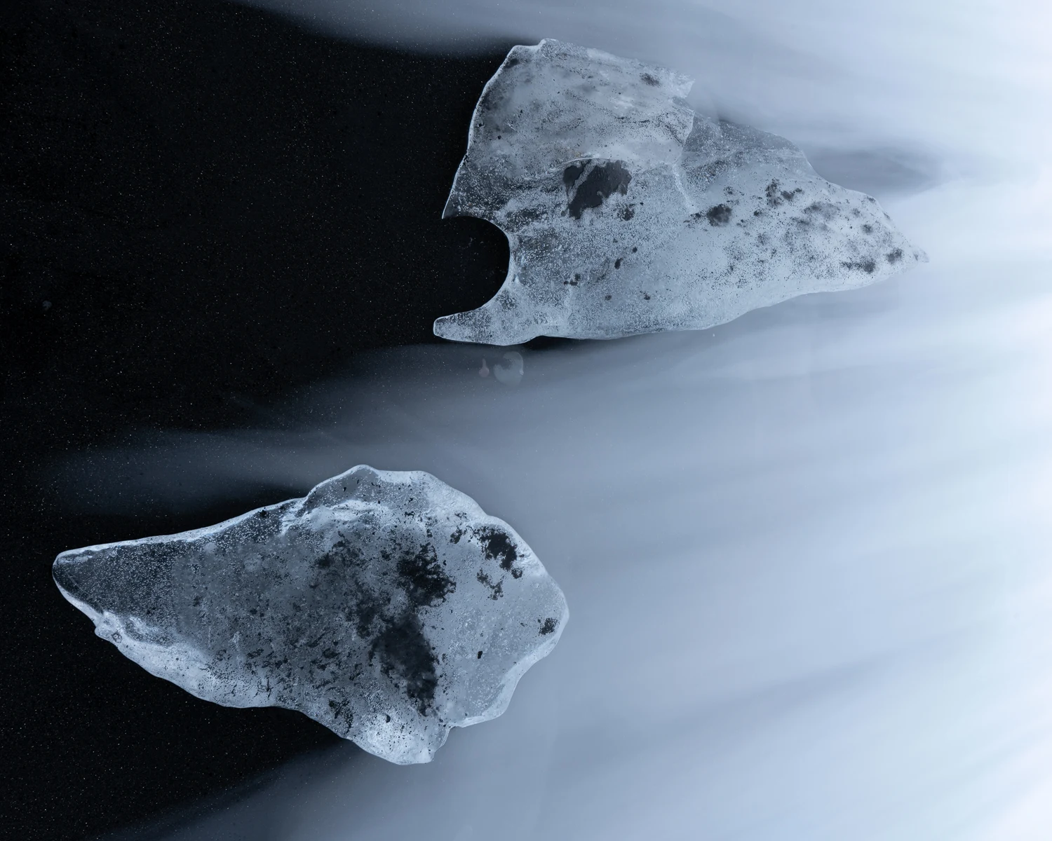 Two irregular pieces of translucent ice rest on black sand, arranged diagonally across the frame as pale, blurred seawater sweeps around their textured surfaces in cool blue-gray tones.