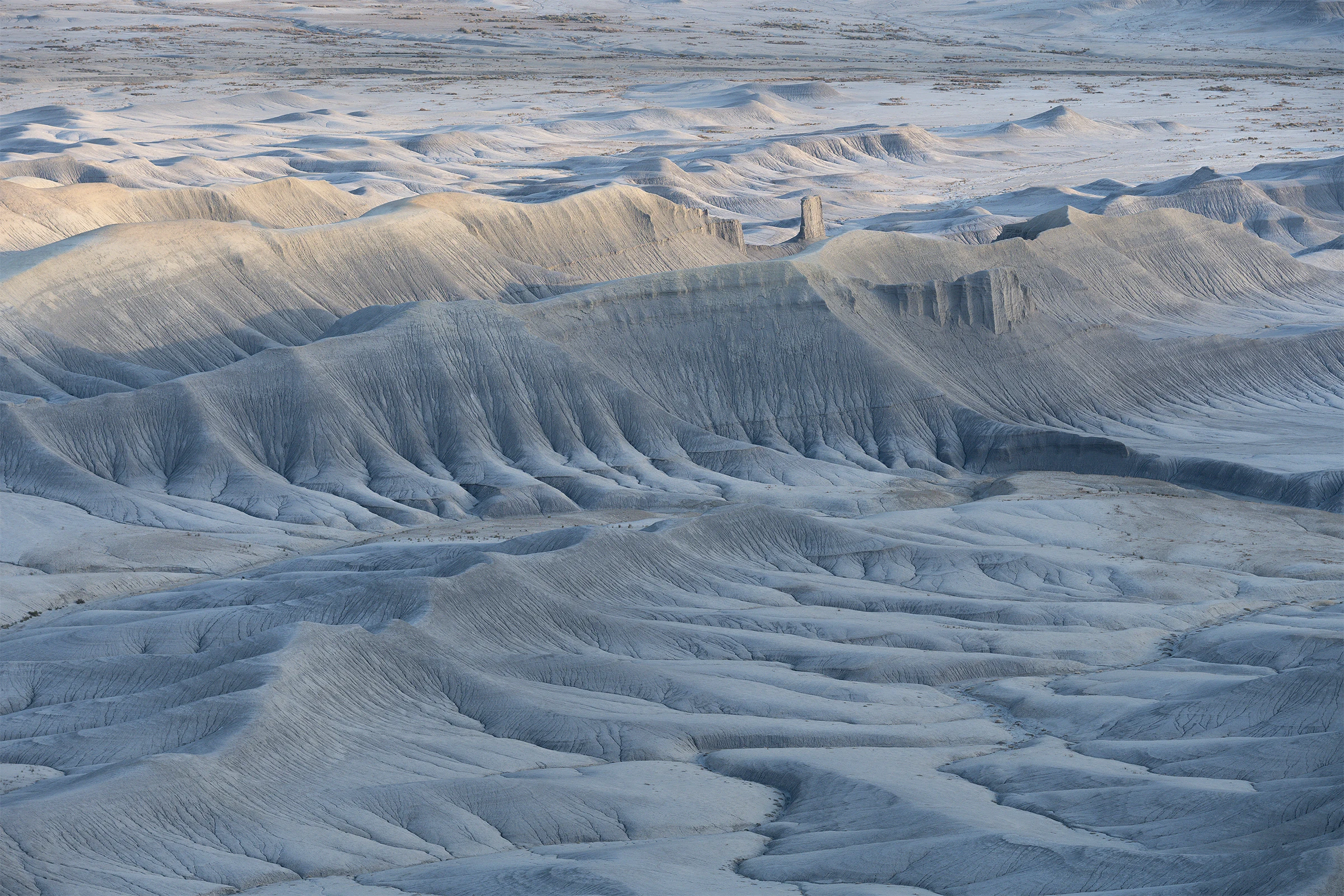 Pale gray badlands form rolling ridges and deeply grooved slopes around winding dry channels, with a narrow rock pillar rising in the middle distance and soft sunlight touching crests above blue-gray shadows.