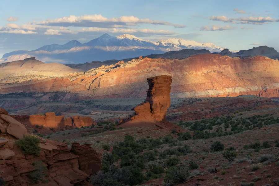 A tall red rock pillar rises above a shrub-dotted desert valley, with layered orange cliffs catching sunlight and snow-covered mountains stretching across the background beneath a blue sky with scattered clouds.