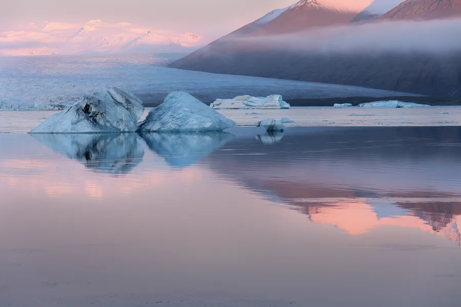 Iceberg reflected in still water beneath pink evening light.