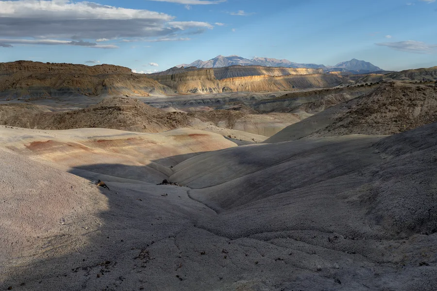 Rounded gray badland hills and eroded gullies lead toward layered cliffs and distant mountains, with patches of golden sunlight and rust-colored bands beneath a partly cloudy blue sky.