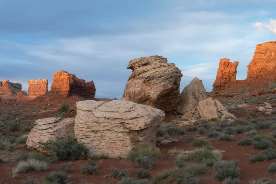 Large pale boulders with tilted layers fill the foreground among scattered desert shrubs, with sunlit red rock towers rising behind them beneath a blue sky streaked with clouds.