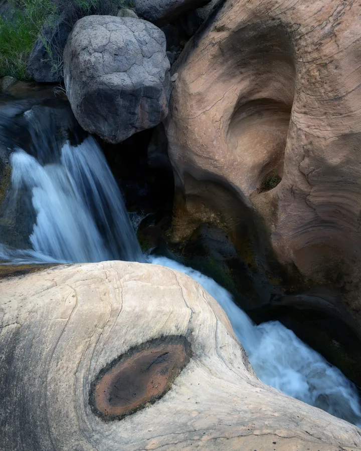 Water cascades between large boulders beside a reddish brown rock wall sculpted with smooth hollows, while pale, veined stone with a shallow pool fills the foreground.