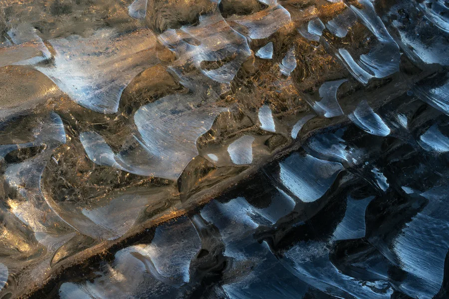 A close-up of translucent ice reveals overlapping curved ridges and scalloped hollows, with warm golden light across the left side contrasting with deep blue reflections and dark crevices on the right.