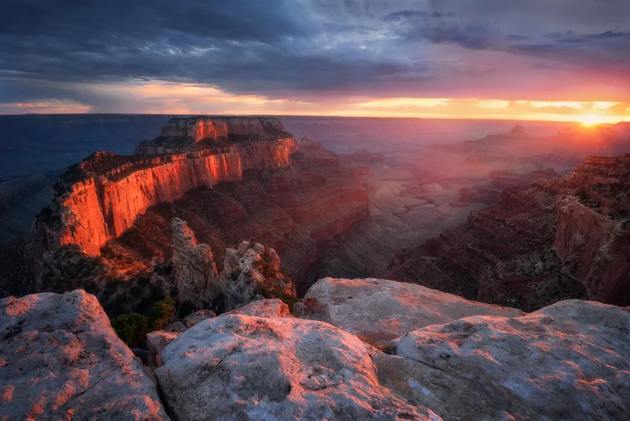 Grand canyon rim at sunset with red cliffs and blue storm clouds.