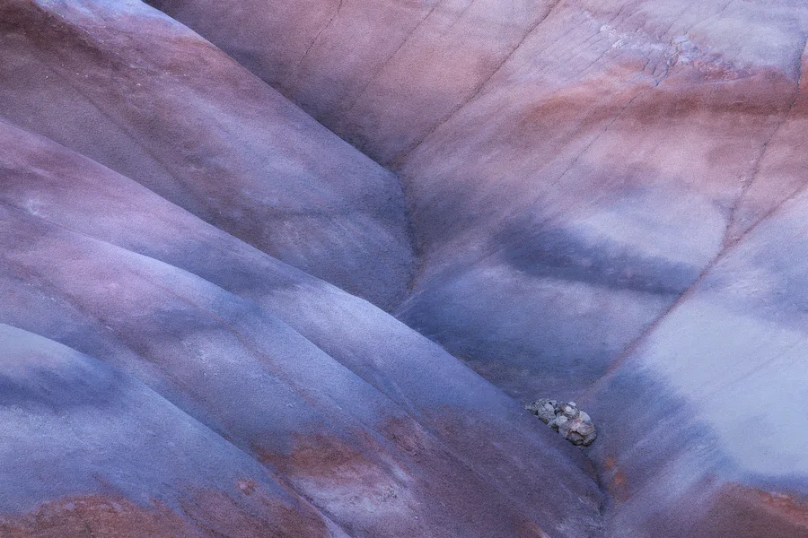 Rounded rock ridges in muted purple, blue, and pink converge around narrow grooves, with soft shadows defining their smooth curves and a small cluster of pale stones lodged near the lower right.