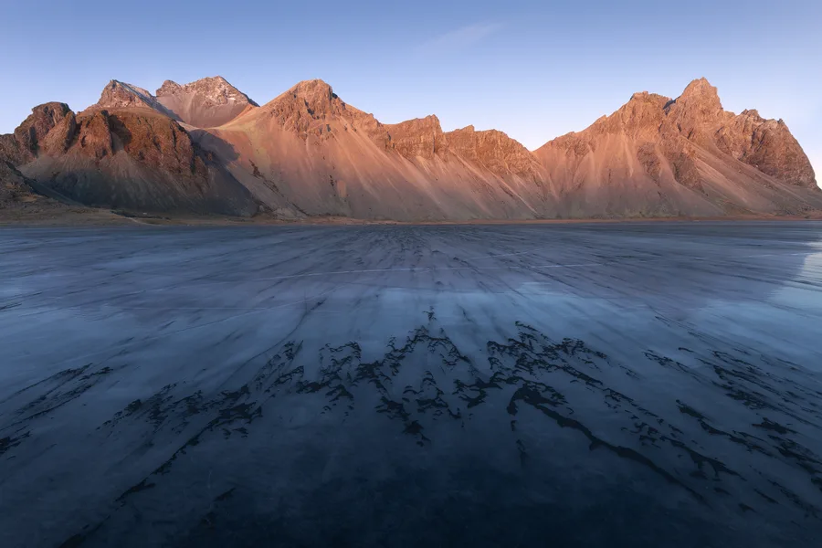 A rugged mountain ridge glows orange above a broad blue-gray expanse of ice, where thin cracks and dark branching streaks lead toward rocky slopes beneath a clear blue sky.
