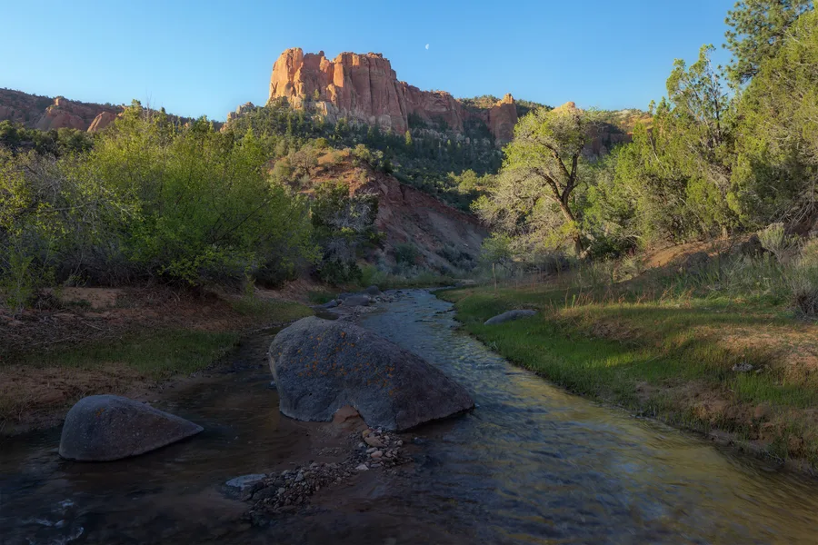 A shallow creek winds around large gray boulders between grassy banks and dense green trees, beneath sunlit red cliffs and a clear blue sky with a small visible moon.