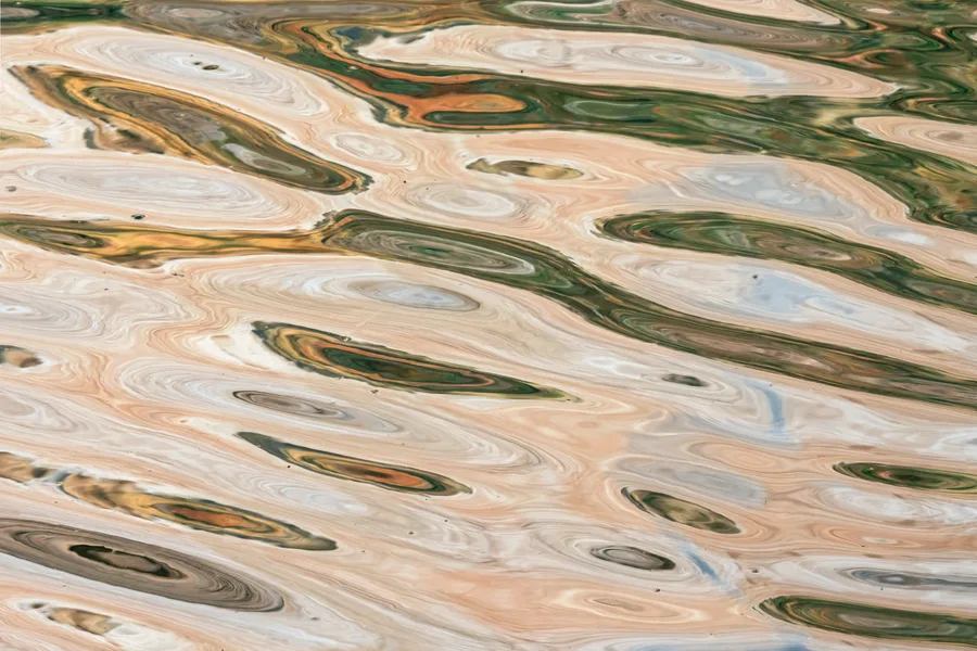 Rippling water distorts cream, peach, gold, and dark green reflections into elongated ovals and flowing bands, creating a tightly framed abstract pattern across the entire surface.