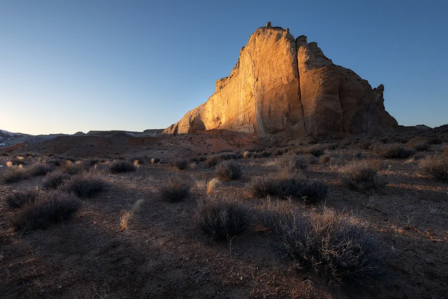 Sunlit desert monolith rising from sagebrush under a blue sky.