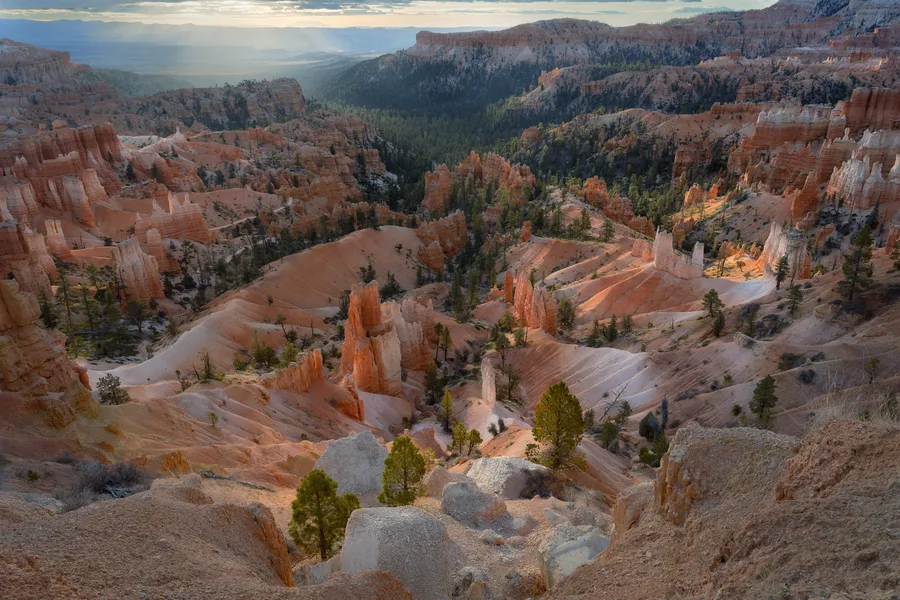 Orange and cream rock spires rise among scattered evergreen trees across a sprawling canyon, viewed from a rocky rim, with sunlight illuminating ridges and hazy distant hills beneath a clouded sky.