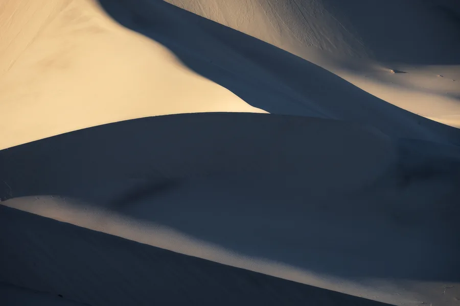 Overlapping sand dunes form sweeping curves, with pale golden sand in sunlight contrasting against deep blue-gray shadows and fine ripples visible along a narrow foreground slope.