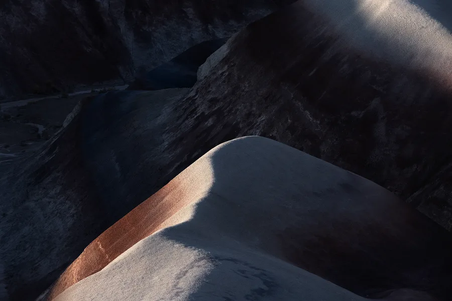 Rounded, barren hills overlap in deep shadow, with narrow bands of sunlight revealing pale gray and rusty red slopes, fine granular textures, and curving ridgelines against darker eroded terrain.