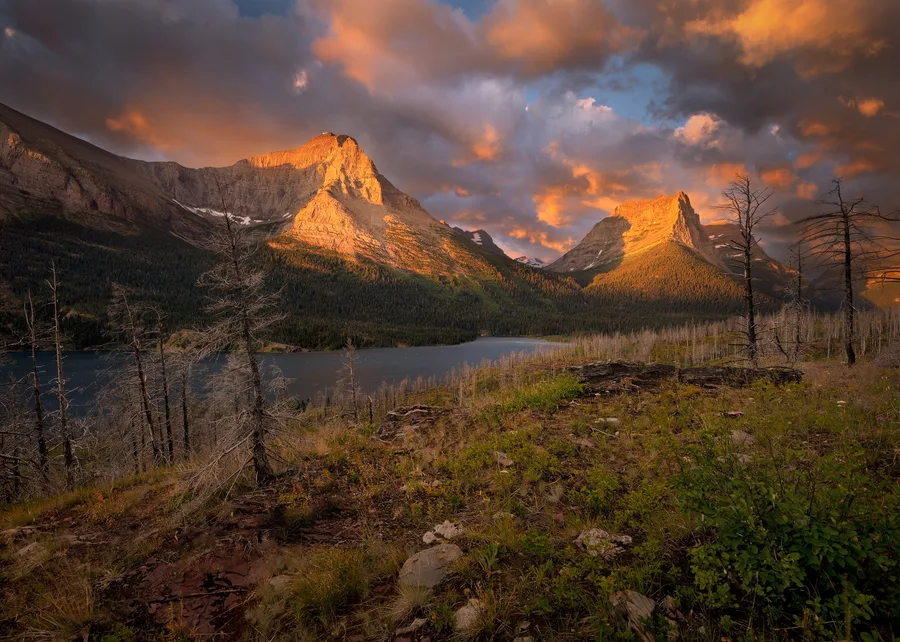 Fiery sunset light breaks across rugged mountain peaks and a dark forest.