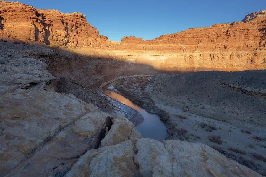 A river curves through a deep canyon beneath layered orange cliffs, with broad rock ledges in the foreground and sparse shrubs along the banks; sunlight illuminates the upper walls above the shadowed canyon floor.