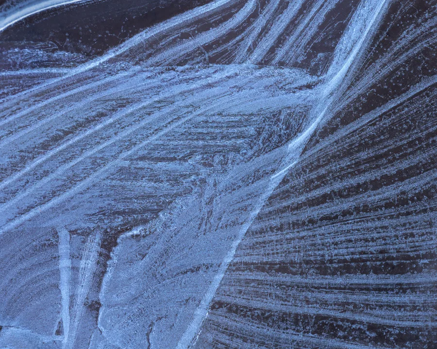 Pale blue ice crystals form overlapping bands across a dark surface, with broad curved streaks intersecting fine, feathery lines and scattered bubbles in a close-up view.