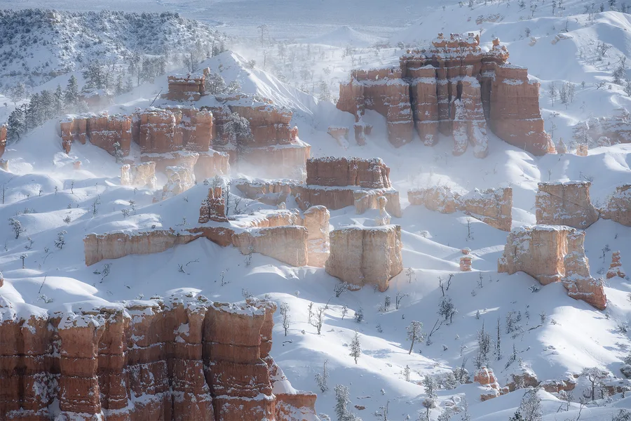 Orange rock towers and layered cliffs rise from deep snow across a canyon, with scattered snow-covered trees, soft blue shadows, and wisps of blowing snow between the formations.