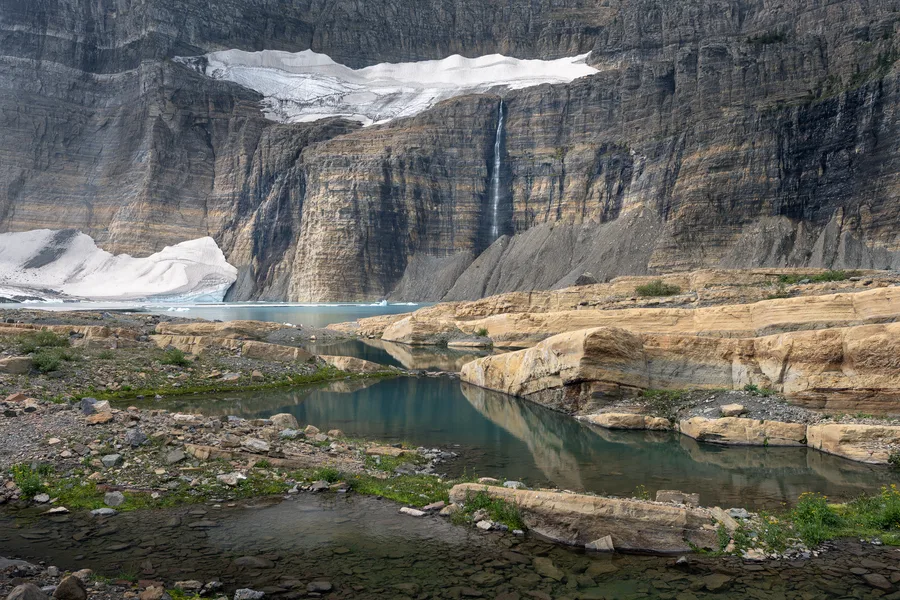 A turquoise glacial lake rests below layered alpine cliffs and snowfields.