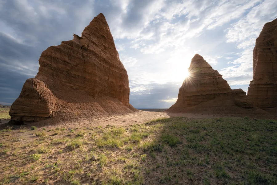 Towering brown rock formations frame a sandy gap, with sparse grasses in the foreground and sunlight bursting around a smaller spire beneath a sky streaked with white and gray clouds.