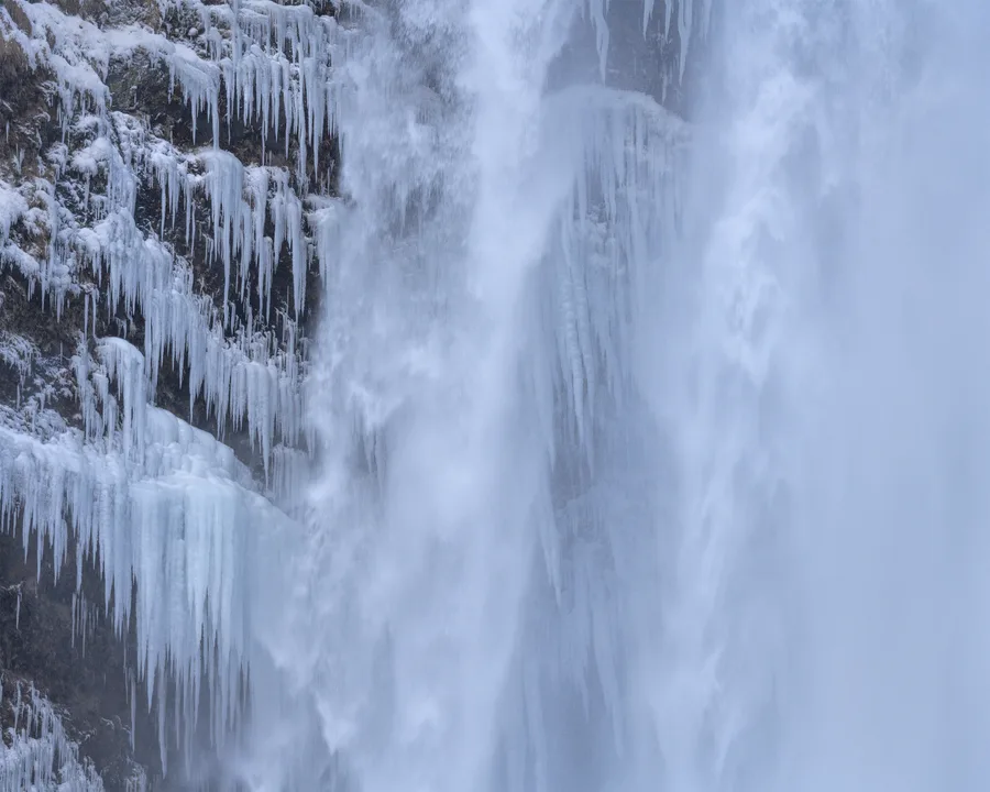 Water cascades beside a dark rock face covered in dense rows of long icicles, with pale blue ice and snow coating the ledges and white spray obscuring much of the right side.