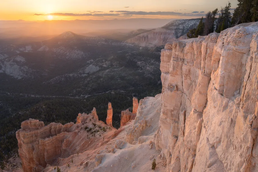 Pale orange cliffs and slender rock spires rise above a forested valley, with a sheer rock face filling the right foreground and a low sun casting golden light across hazy distant ridges.