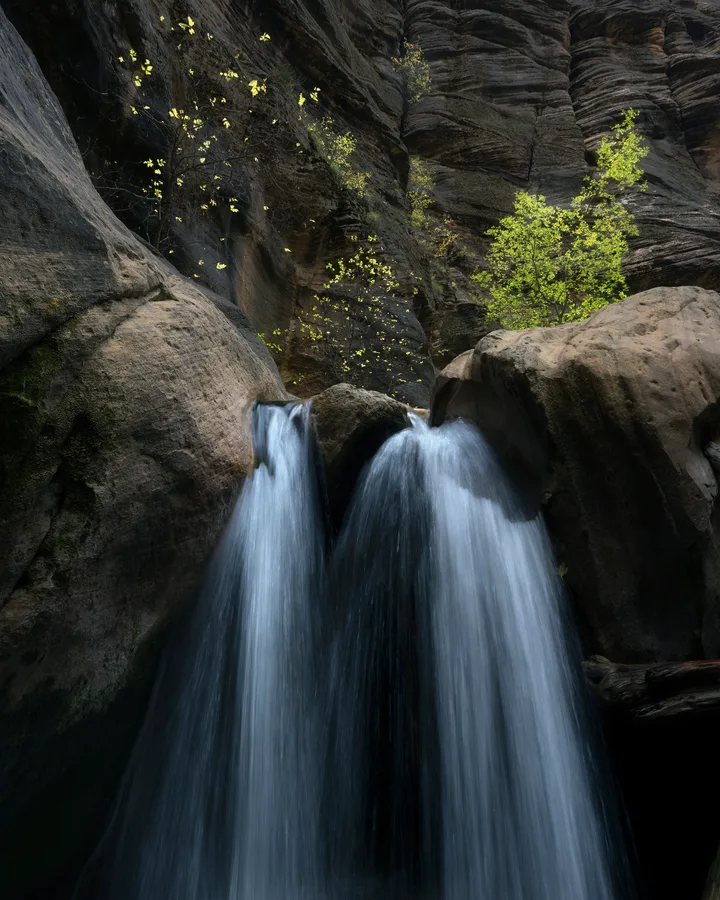 A waterfall splits around a central boulder and drops between large rocks beneath towering, layered canyon walls, with sunlit green leaves standing out against the shaded stone.