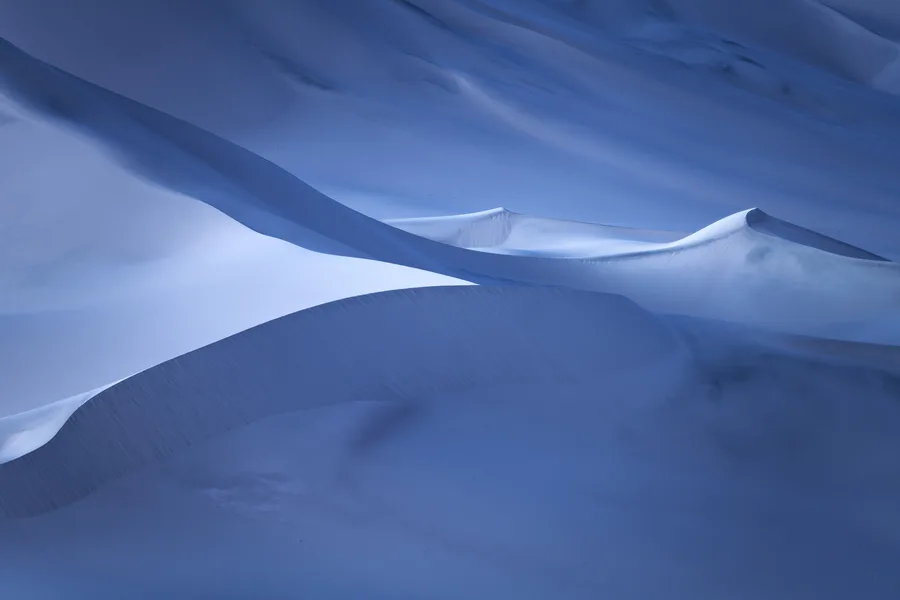 Overlapping sand dunes form sweeping curves and sharp crests, with pale illuminated edges contrasting against deep blue shadows and fine ripples texturing the steep foreground slope.