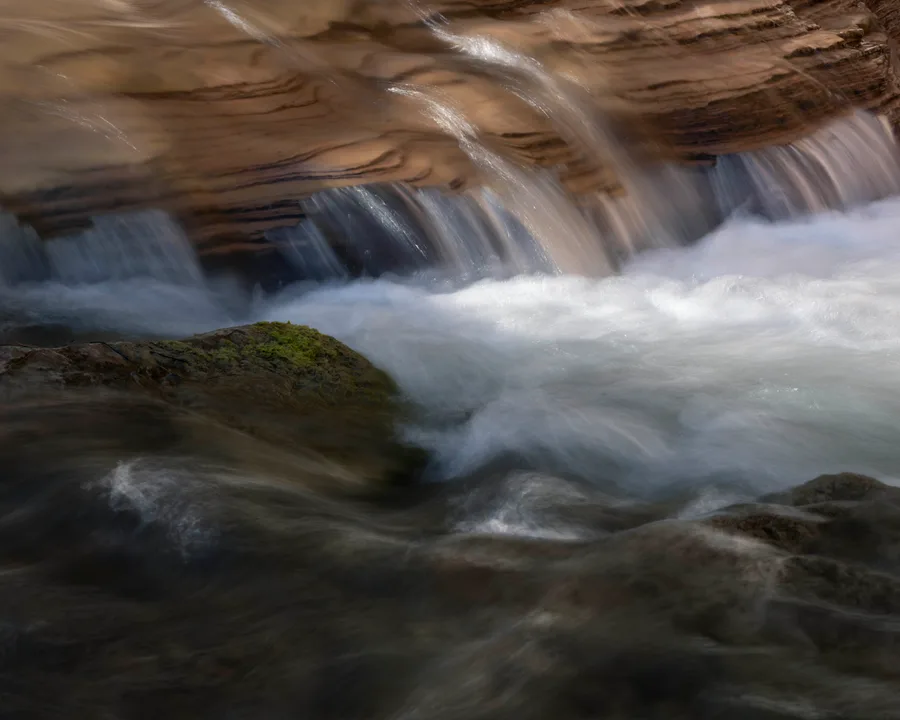 Water cascades over layered brown rock ledges into white rapids, flowing around a moss-covered rock and across dark foreground stones in soft streaks of motion.