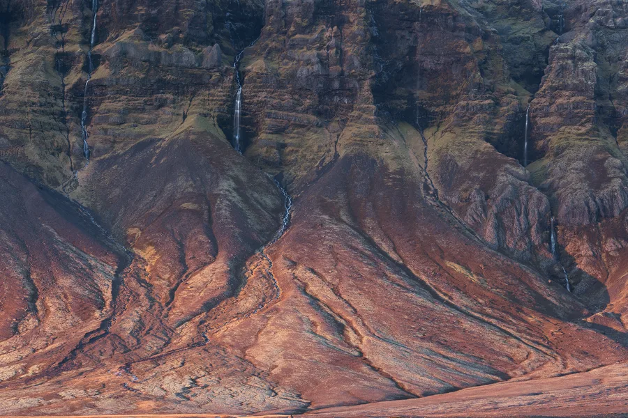 Thin waterfalls descend through dark, layered cliffs into winding streams across steep, rust-brown slopes, where patches of green vegetation and deep shadows emphasize ridges and branching erosion channels.