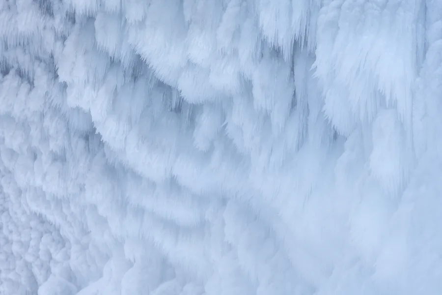 Dense layers of pale blue and white ice cover a vertical surface, with slender icicles hanging beneath rounded, feathery formations and soft blue shadows defining the overlapping ridges.