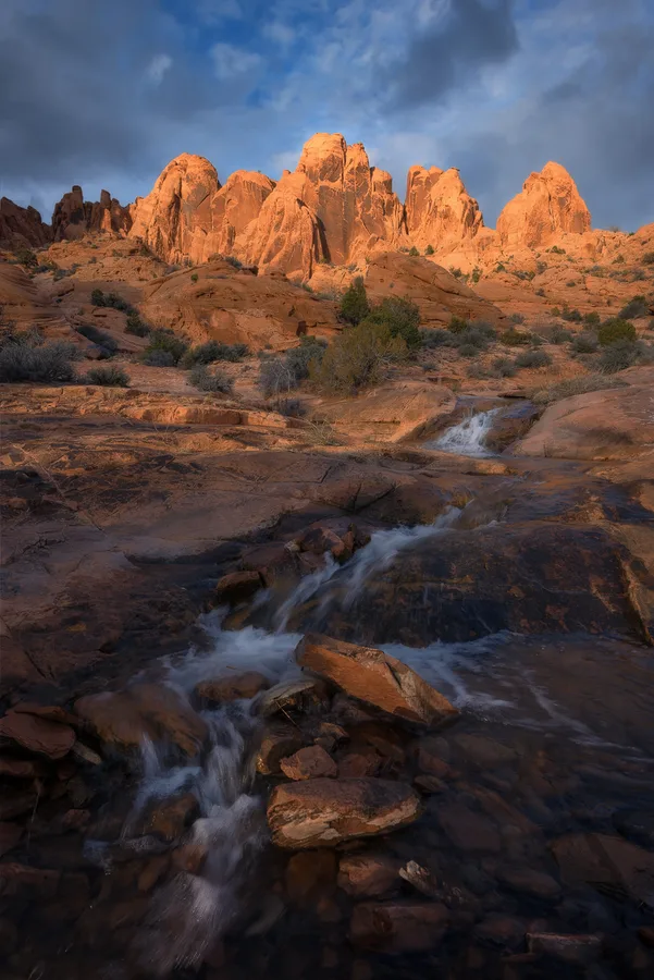 Desert stream beneath glowing red rock and blue storm clouds.