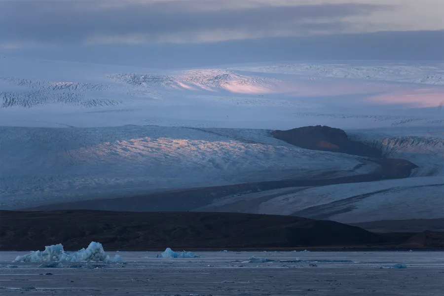 A broad, crevassed glacier rises behind a dark rocky ridge and a lagoon scattered with blue icebergs, with soft pink light touching the snow beneath low gray clouds.