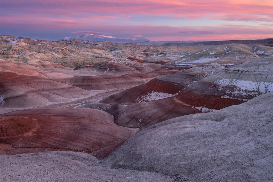 Rounded badlands striped with red and gray layers stretch toward distant snow-covered mountains, with scattered snow tracing eroded slopes beneath a broad sky of pink and purple clouds.