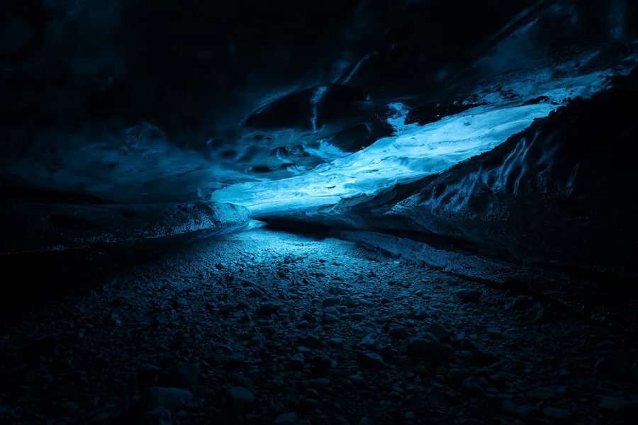 A low ice cave curves above a gravel-covered floor, with a luminous blue band crossing the sculpted ceiling diagonally and casting blue light onto stones between dark walls.