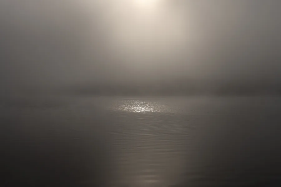 A pale sun glows through dense fog above still water.