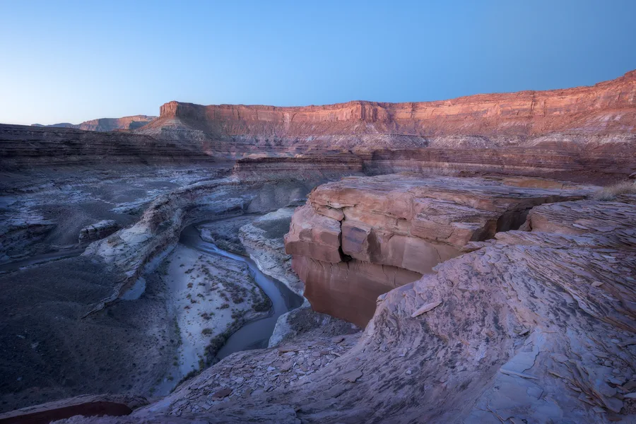A narrow river winds through a deep, layered canyon below a broad sandstone ledge, with pink light on distant red cliffs and cool shadows across the rocky foreground beneath a clear blue sky.