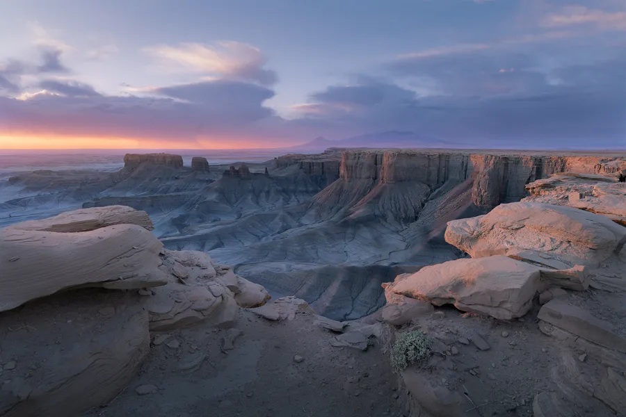 Flat-topped cliffs and deeply eroded desert ridges stretch beyond layered foreground rocks, with distant mountains beneath blue-gray clouds and a pink-orange glow along the horizon.