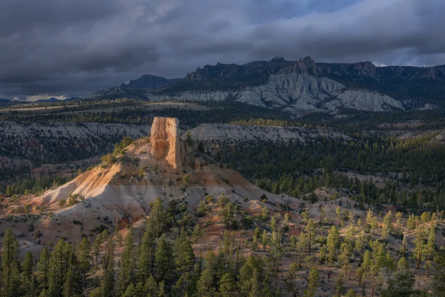 A sunlit orange rock tower rises above pale, eroded slopes dotted with evergreen trees, with forested ridges and rugged cliffs stretching beneath dark clouds in the background.