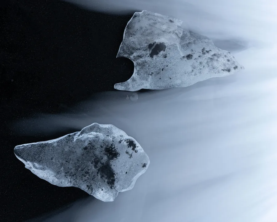 Two irregular pieces of translucent ice rest on black sand, arranged diagonally across the frame as pale, blurred seawater sweeps around their textured surfaces in cool blue-gray tones.