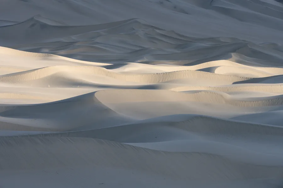 Overlapping sand dunes fill the frame, with a band of pale golden sunlight across the middle contrasting with blue-gray shadows and revealing fine ripples along curved slopes and ridges.